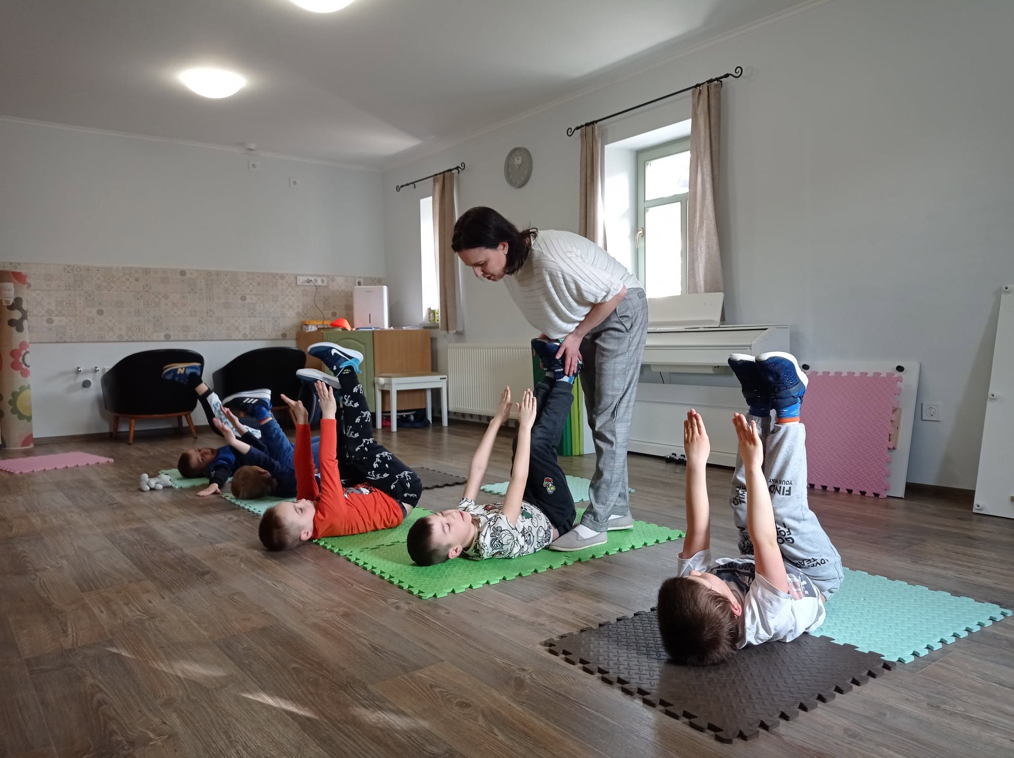 Terapeuta szőnyegen hanyatt fekvő gyerekek lábemelő gyakorlatát segíti