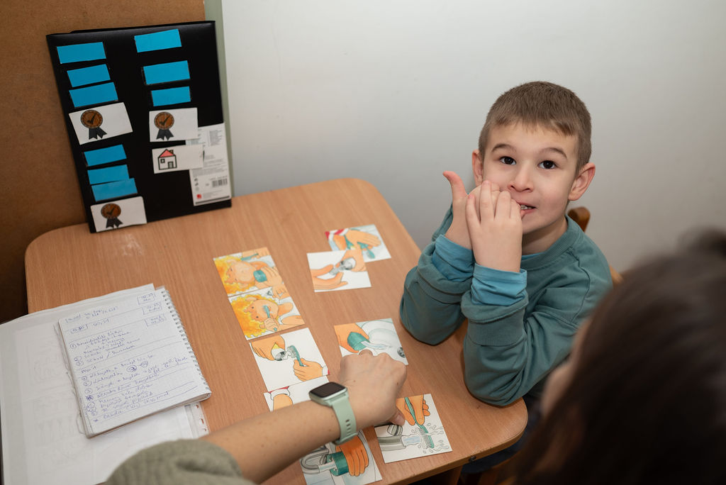 Kisfiú ajakgyakorlatot mutat a logopédussal képkártyákkal teli asztalnál