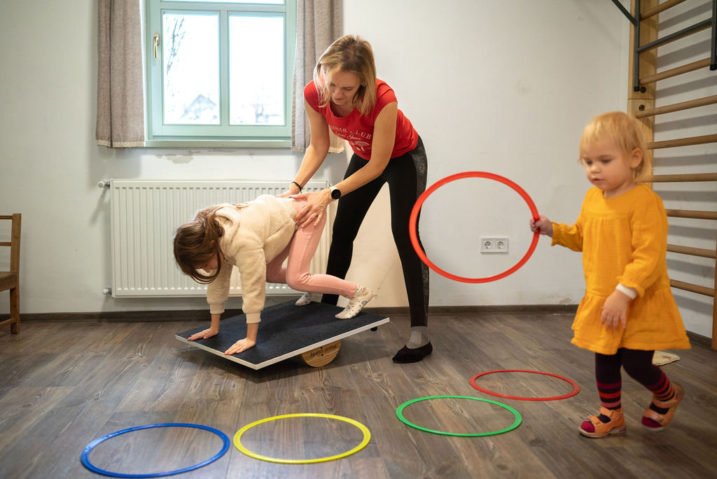 Terapeuta billenődeszkán támogat egy kislányt, mellette karikát tartó kisgyermek